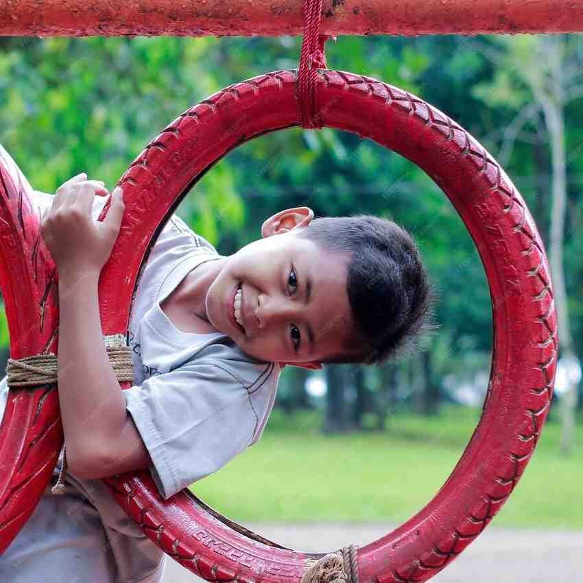 A boy smiling and hanging from a red tire swing in a playground.