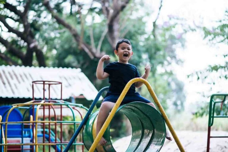 A young boy in a black shirt excitedly sits on a green slide at a playground.